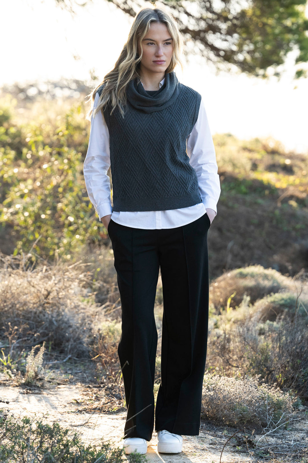 Woman wearing a dark gray sleeveless sweater over a white shirt with black pants in a natural setting.