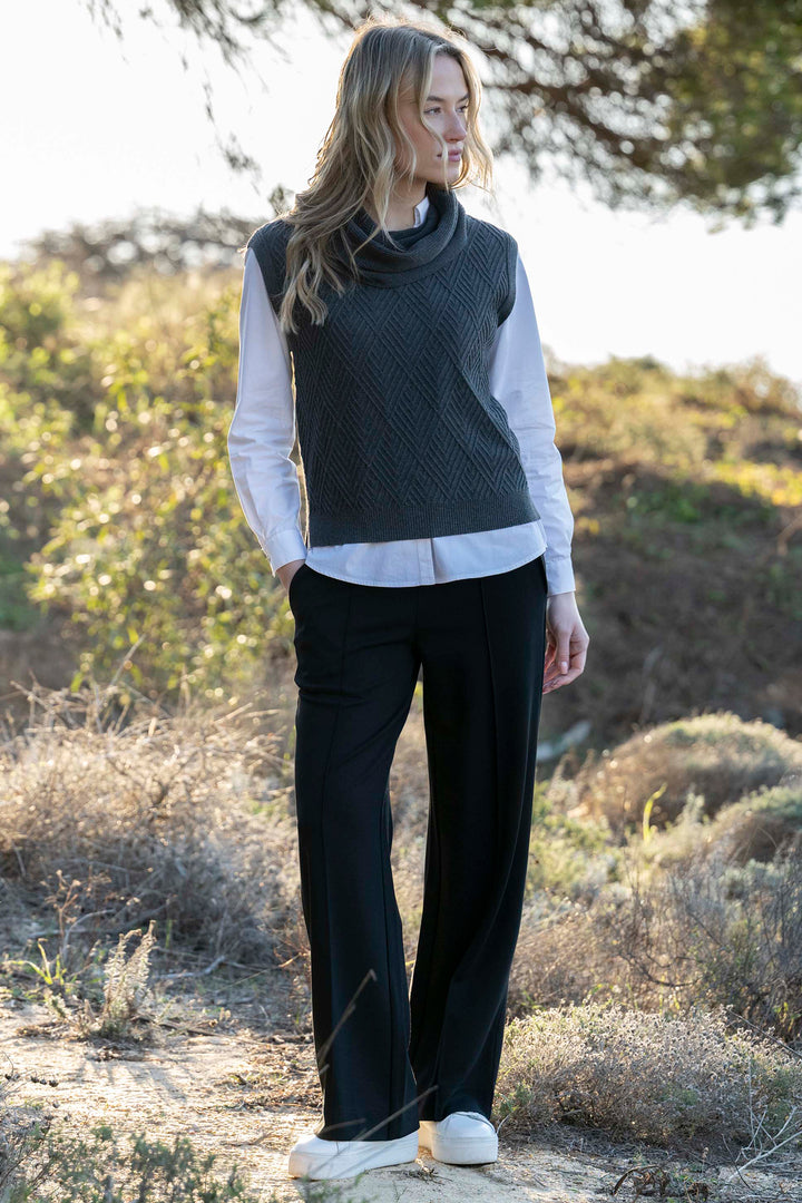 Woman wearing a dark gray sleeveless sweater over a white shirt with black pants in a natural setting.