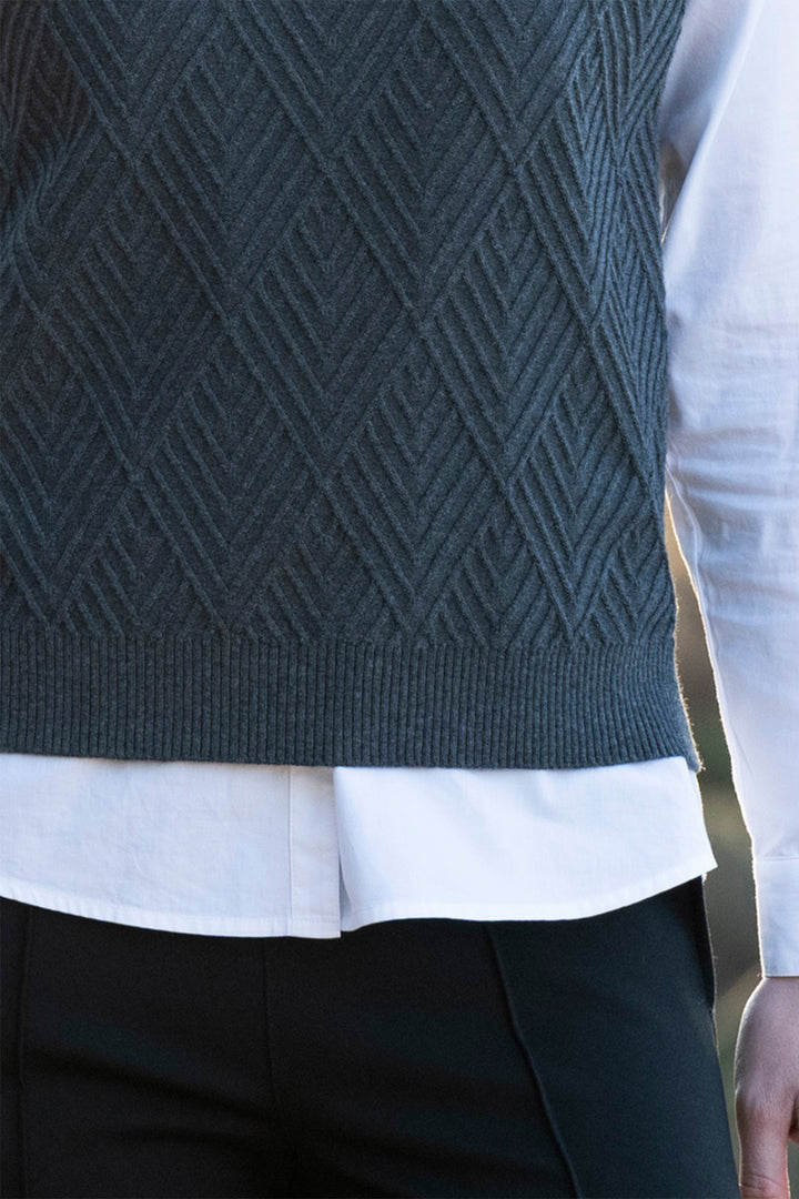 Person wearing a dark green patterned sweater vest over a white shirt and black pants.