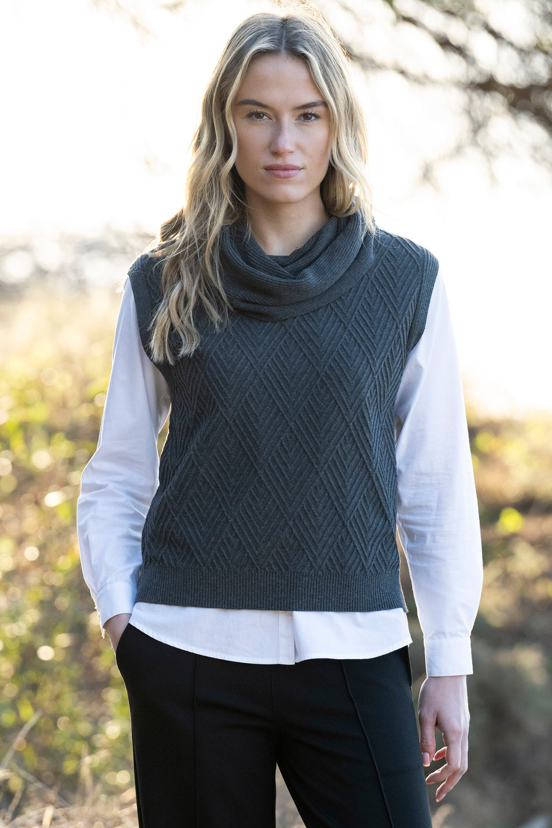 Woman wearing a dark green knitted vest over a white shirt with a blurred natural background