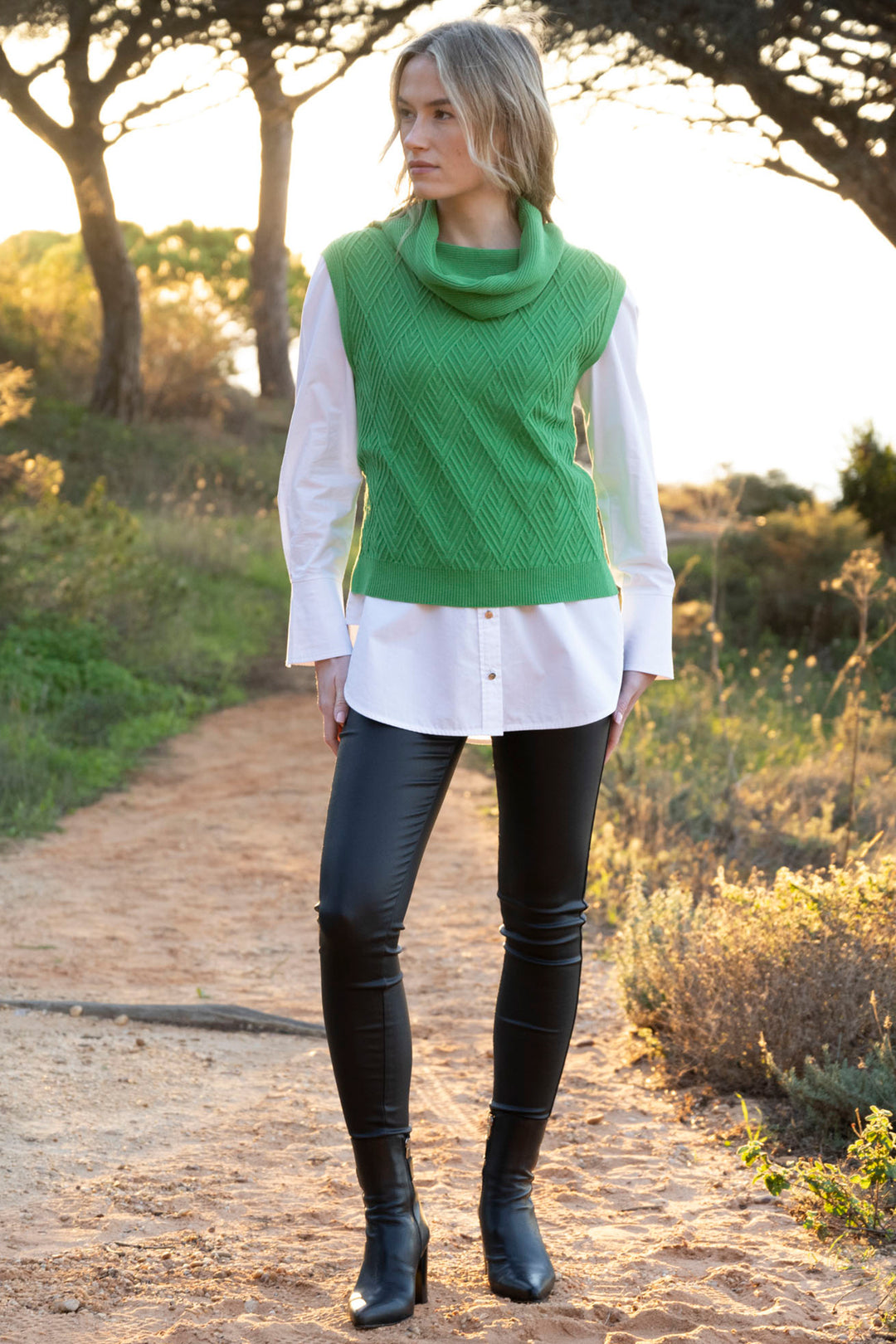 Woman wearing a green sleeveless sweater vest over a white shirt with black pants and boots, standing on a dirt path.