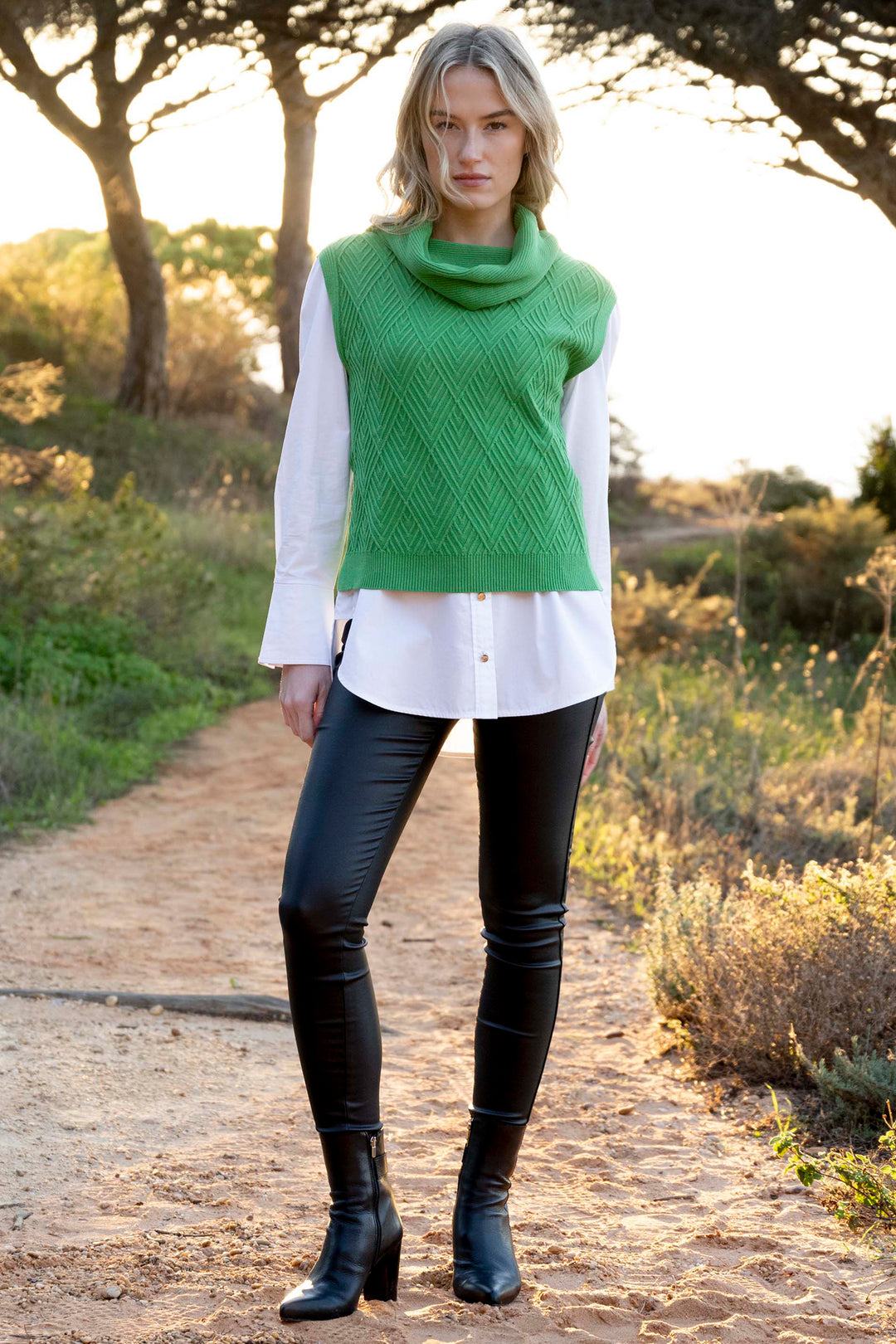Woman wearing a green sleeveless sweater, white shirt, black pants, and black boots standing on a path with trees and grass in the background.