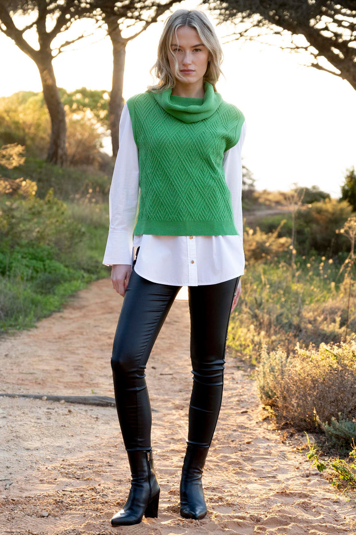 Woman wearing a green sleeveless sweater, white shirt, black pants, and black boots standing on a path with trees and grass in the background.