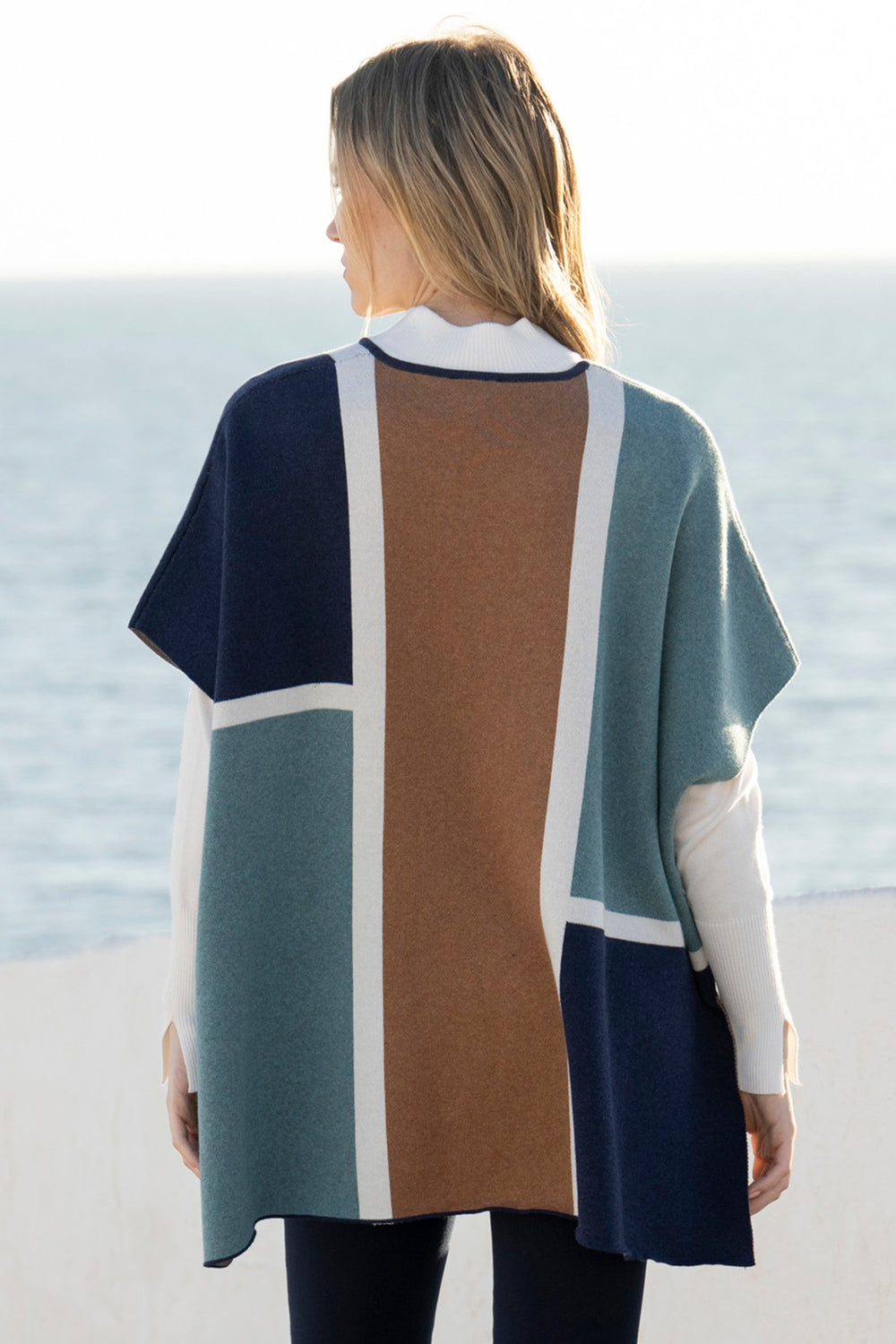 Person wearing a color-blocked poncho with a pattern of blue, green, brown, and white, standing on a beach.