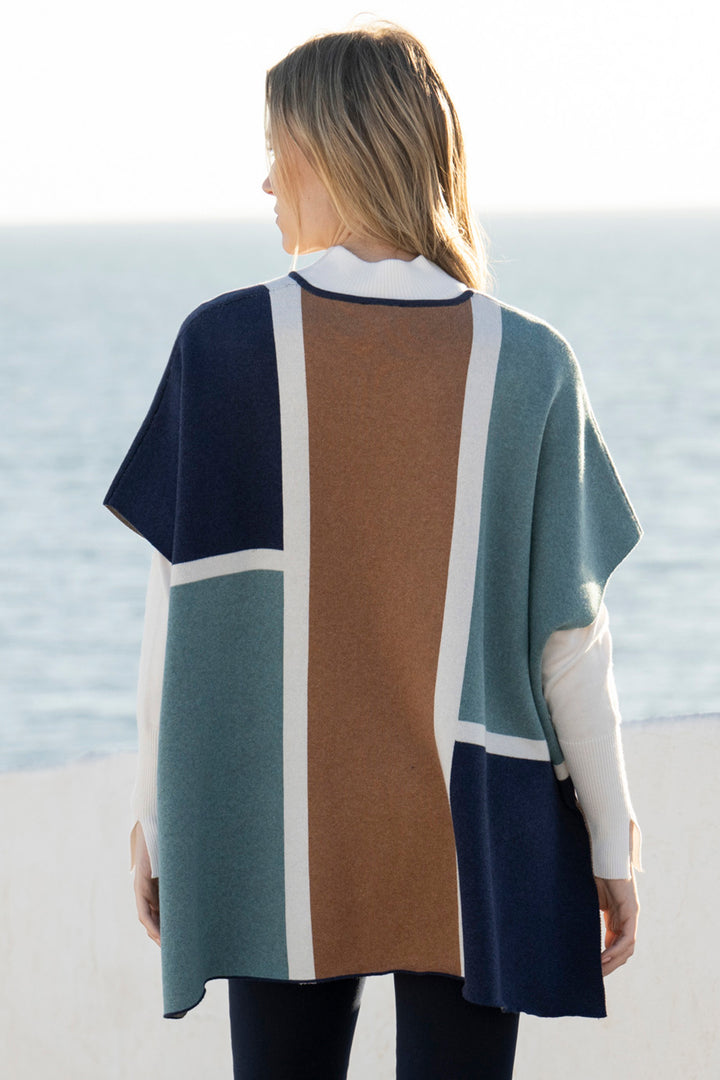 Person wearing a color-blocked poncho with a pattern of blue, green, brown, and white, standing on a beach.