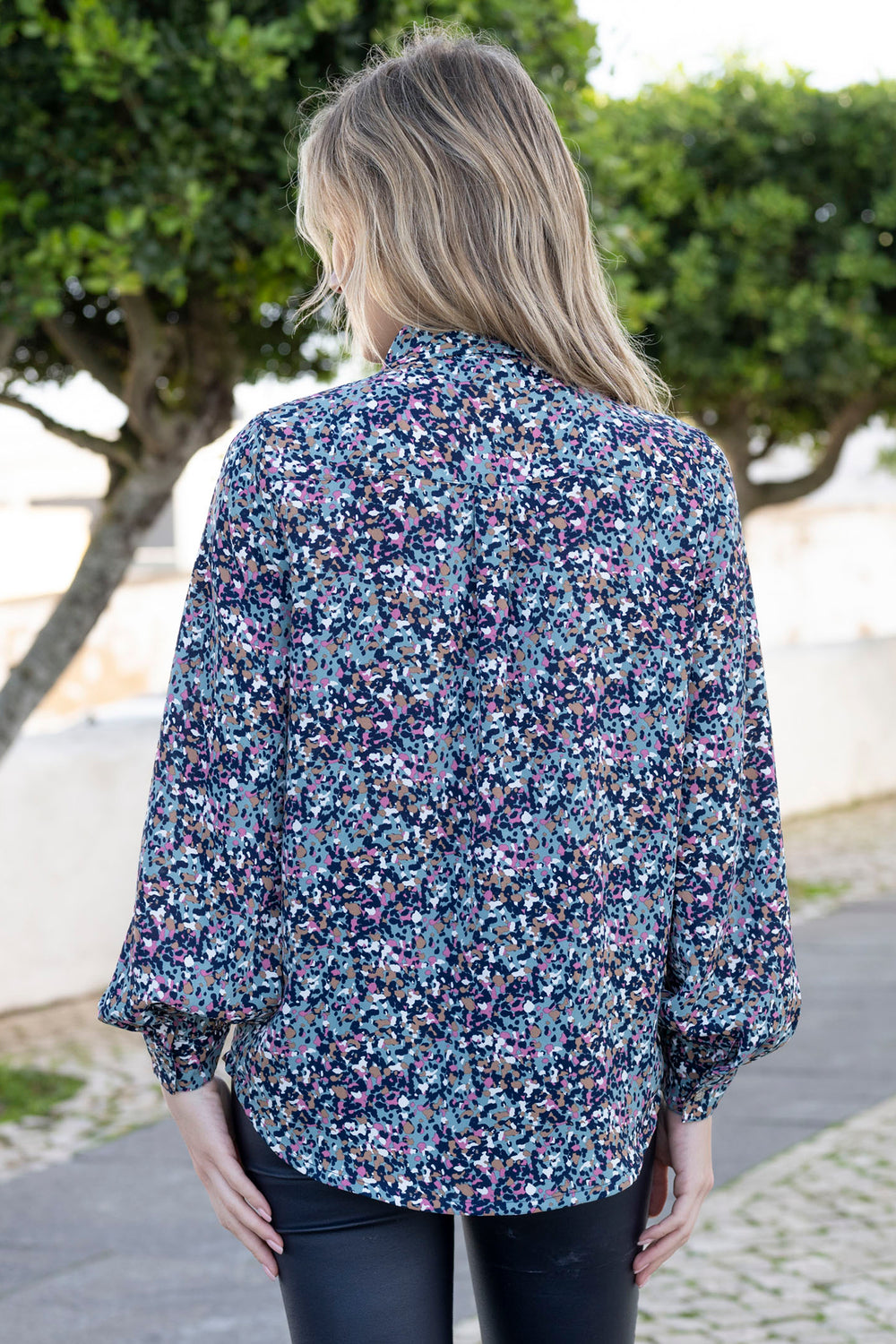 Woman wearing a floral blouse with a blurred background of greenery and a path.