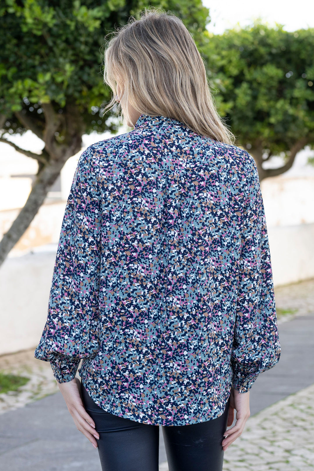 Woman wearing a floral blouse with a blurred background of greenery and a path.