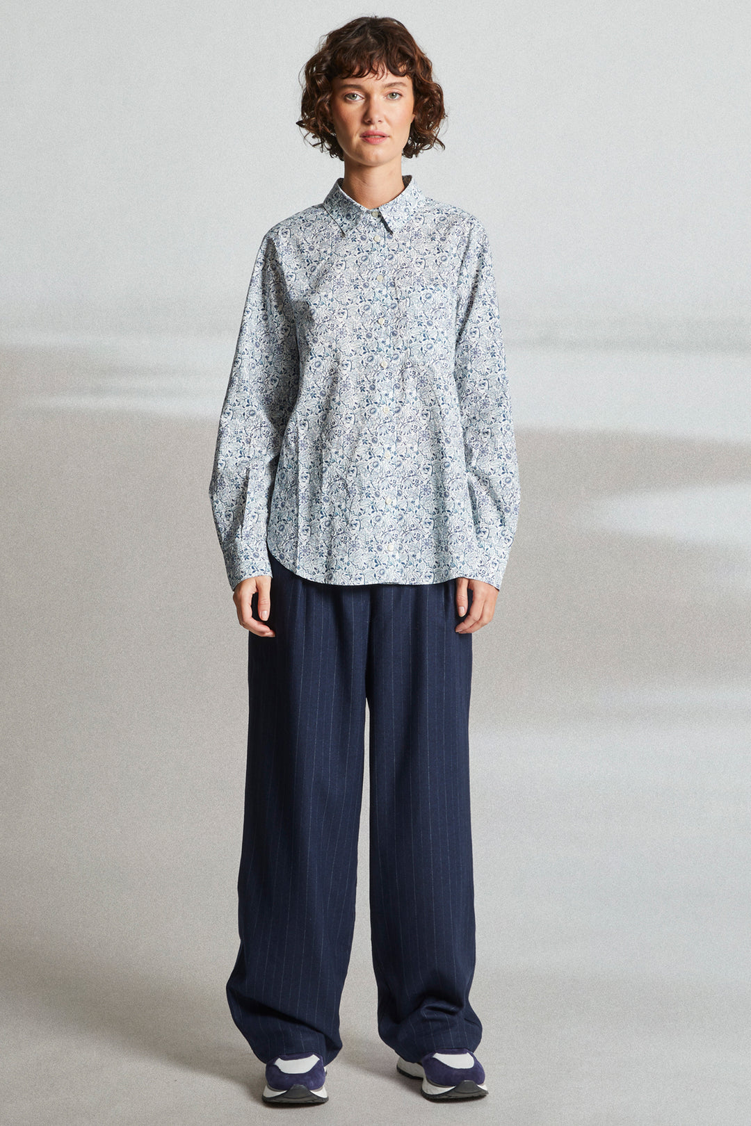 Person wearing a light patterned shirt and dark pants on a plain background