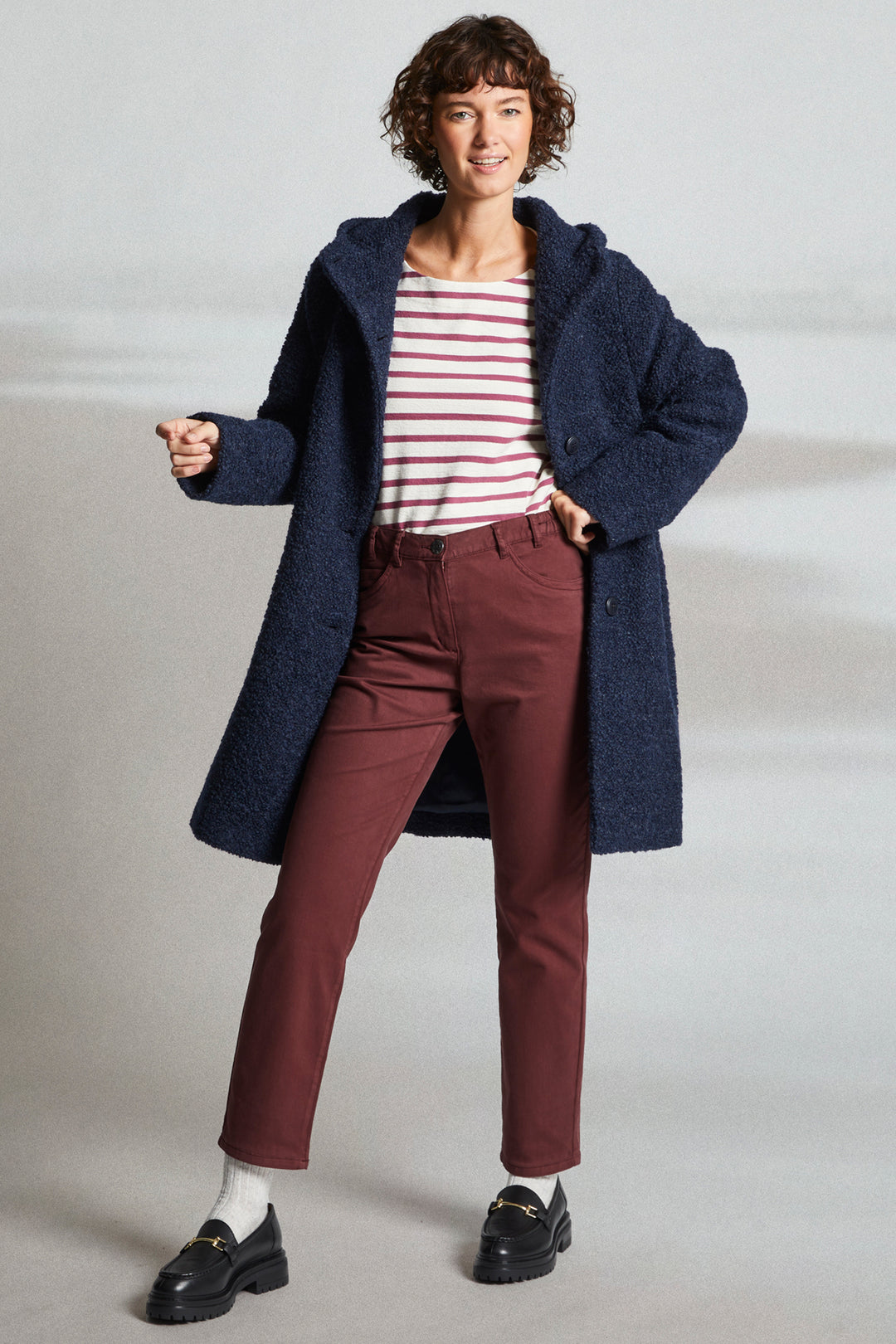 Woman wearing a long navy coat, striped shirt, and maroon pants on a light background