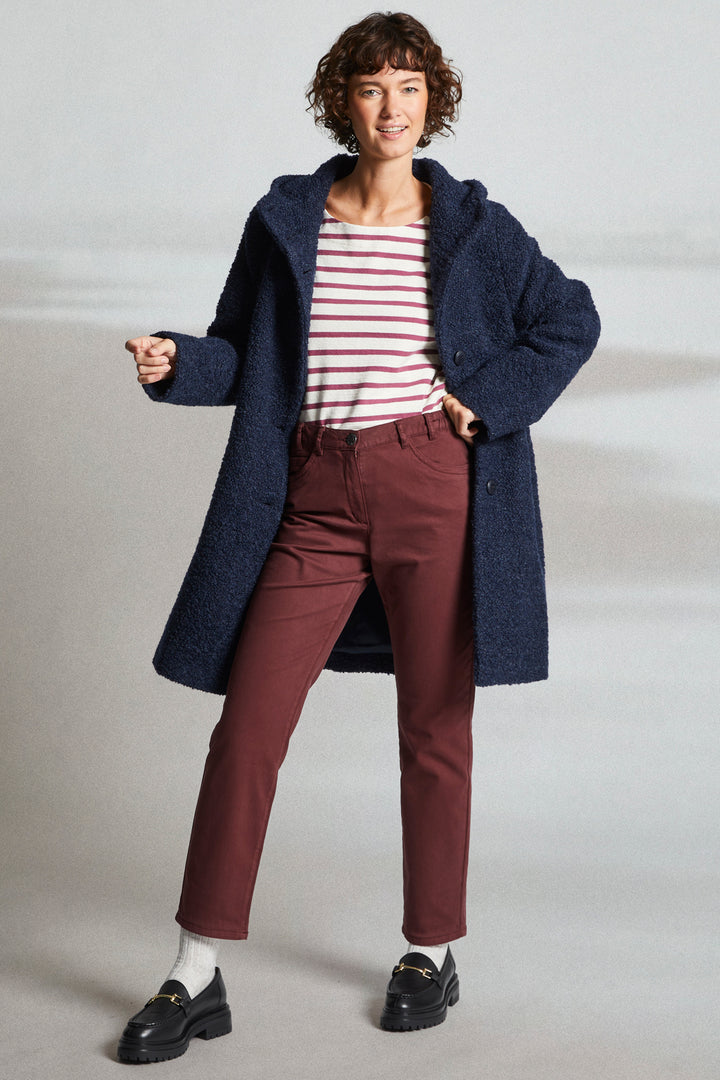 Woman wearing a long navy coat, striped shirt, and maroon pants on a light background
