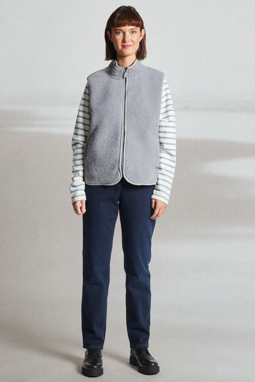 Person wearing a gray quilted vest over a striped shirt with navy pants on a plain background
