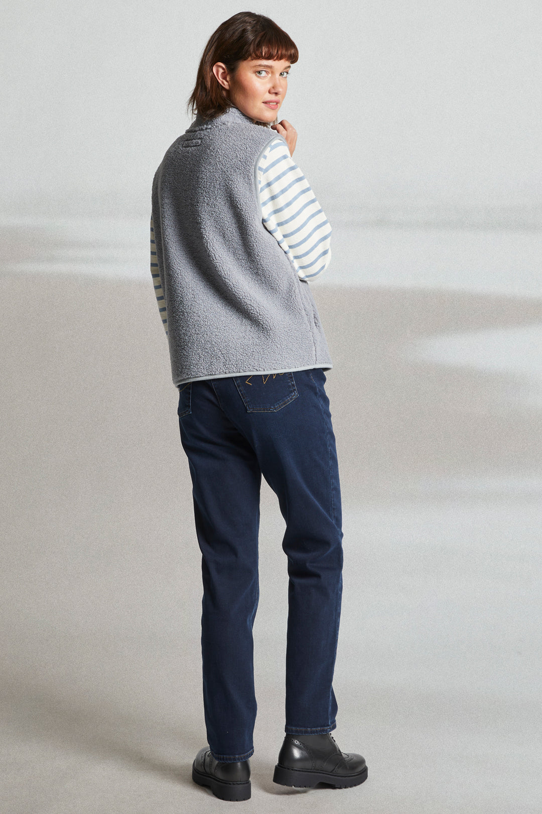 Person wearing a gray sweater over a striped shirt with blue jeans on a plain background