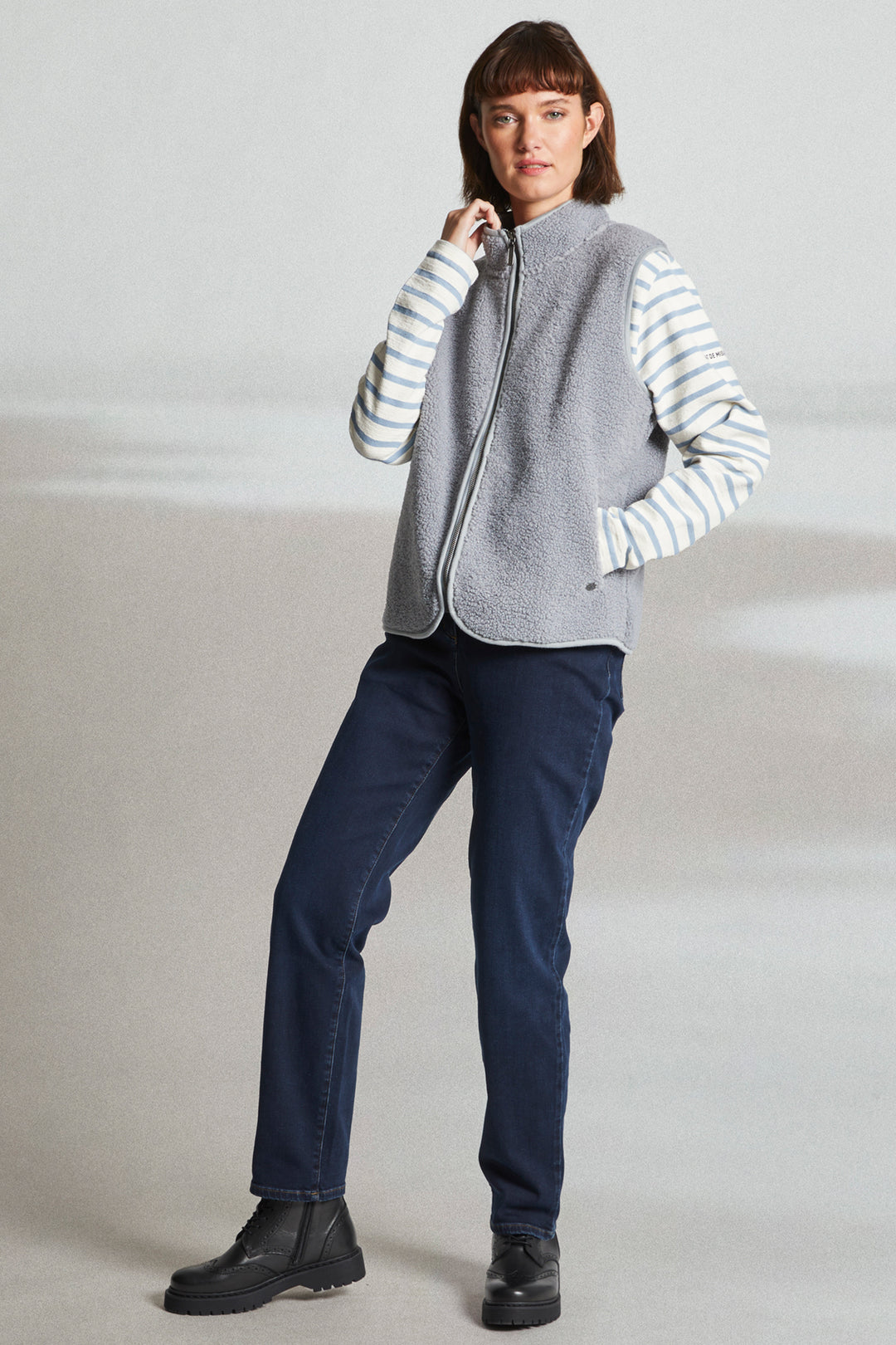 Woman wearing a gray zip-up vest over a striped shirt with navy pants on a plain background