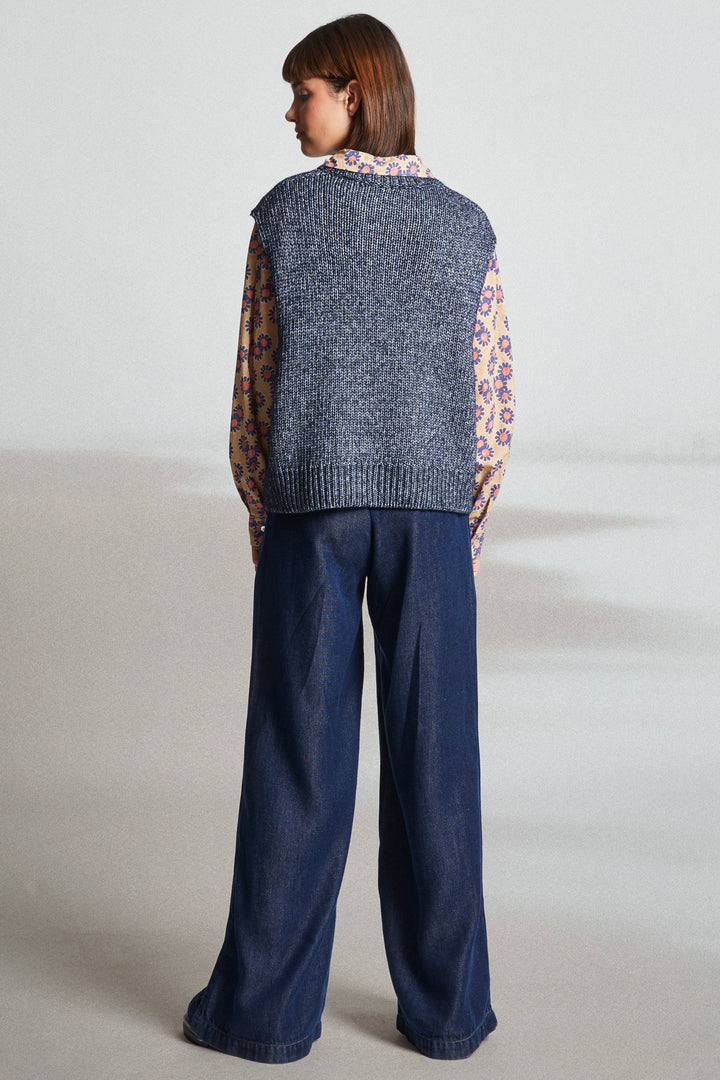 Person wearing a blue knitted vest and dark blue pants on a plain background