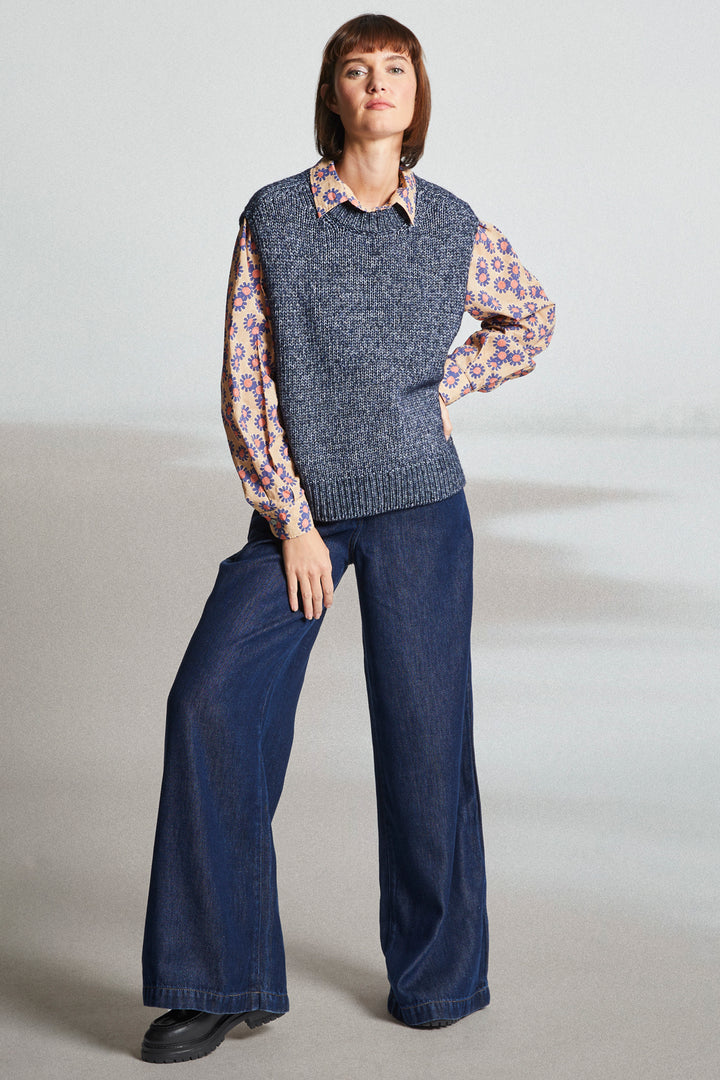 Woman wearing a blue knitted vest over a floral shirt with wide-leg jeans on a plain background