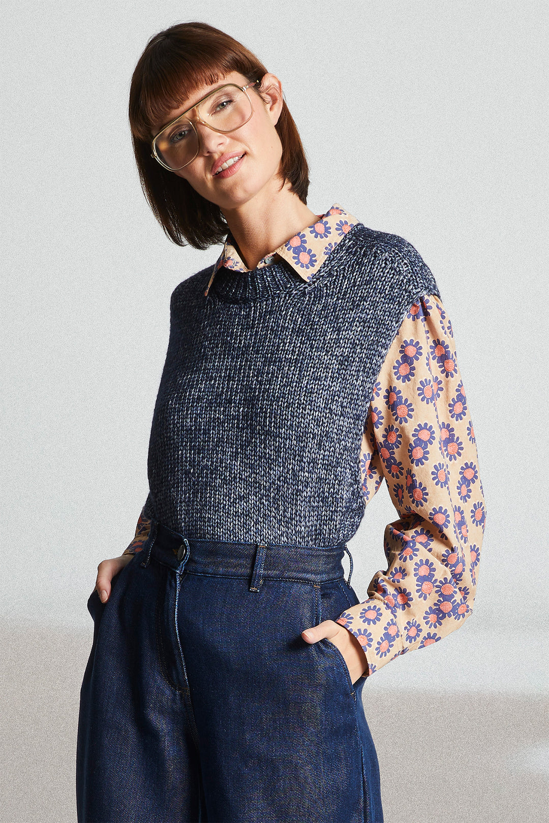 Woman wearing a dark blue knitted sweater over a patterned shirt with denim pants on a light background