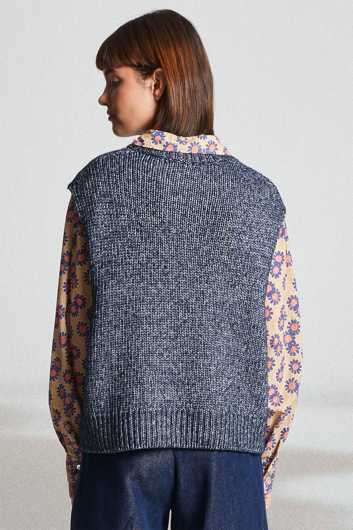 Woman wearing a gray knitted vest over a colorful floral shirt with a plain background