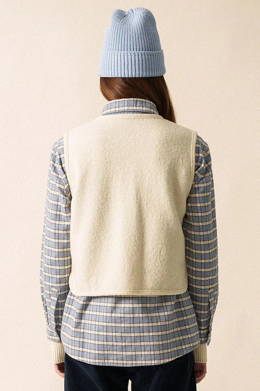 Person wearing a light blue knit hat, cream vest, and plaid shirt on a beige background