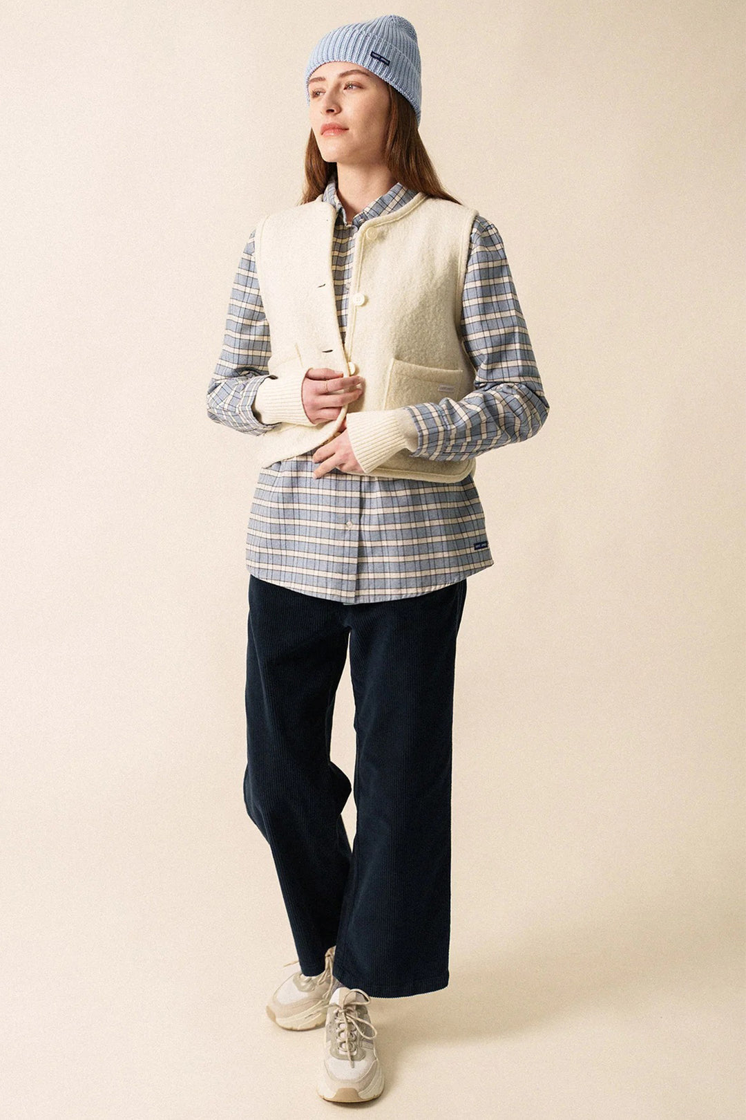 Person wearing a plaid shirt, cream vest, navy pants, and white sneakers on a beige background