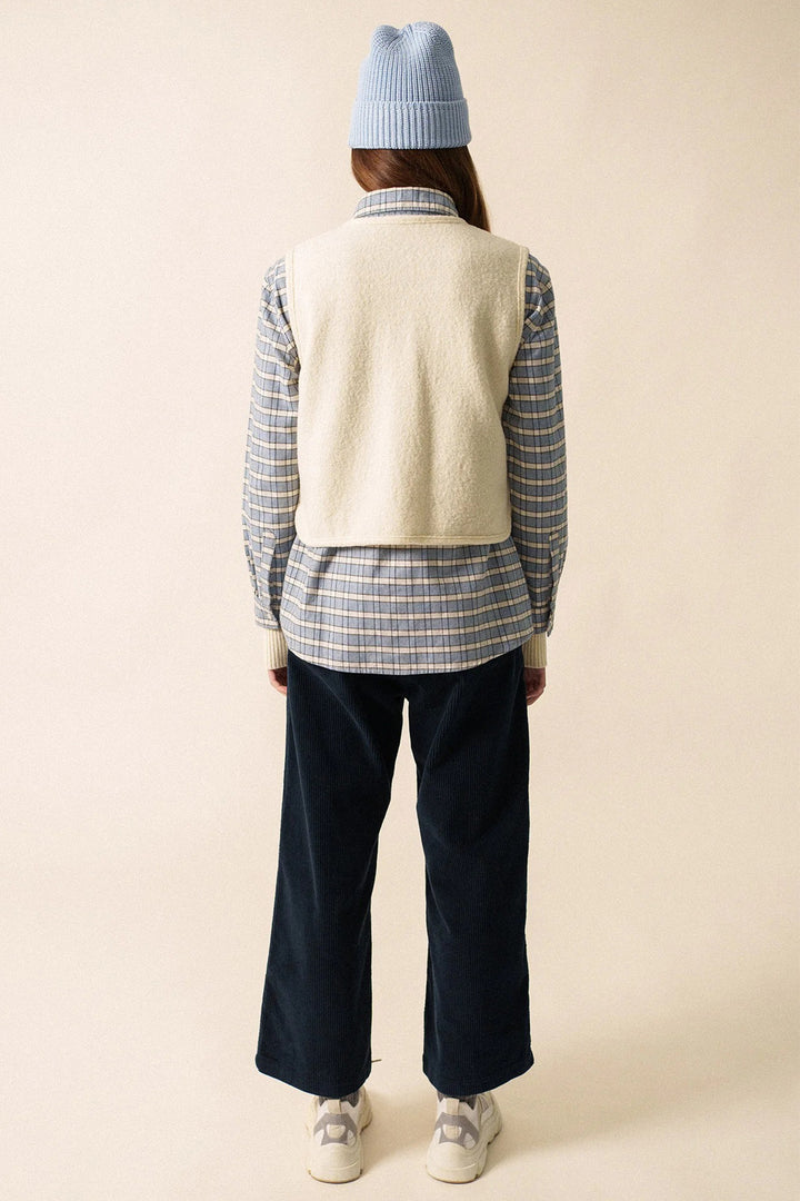 Person wearing a plaid shirt, cream vest, blue pants, and a blue beanie on a beige background