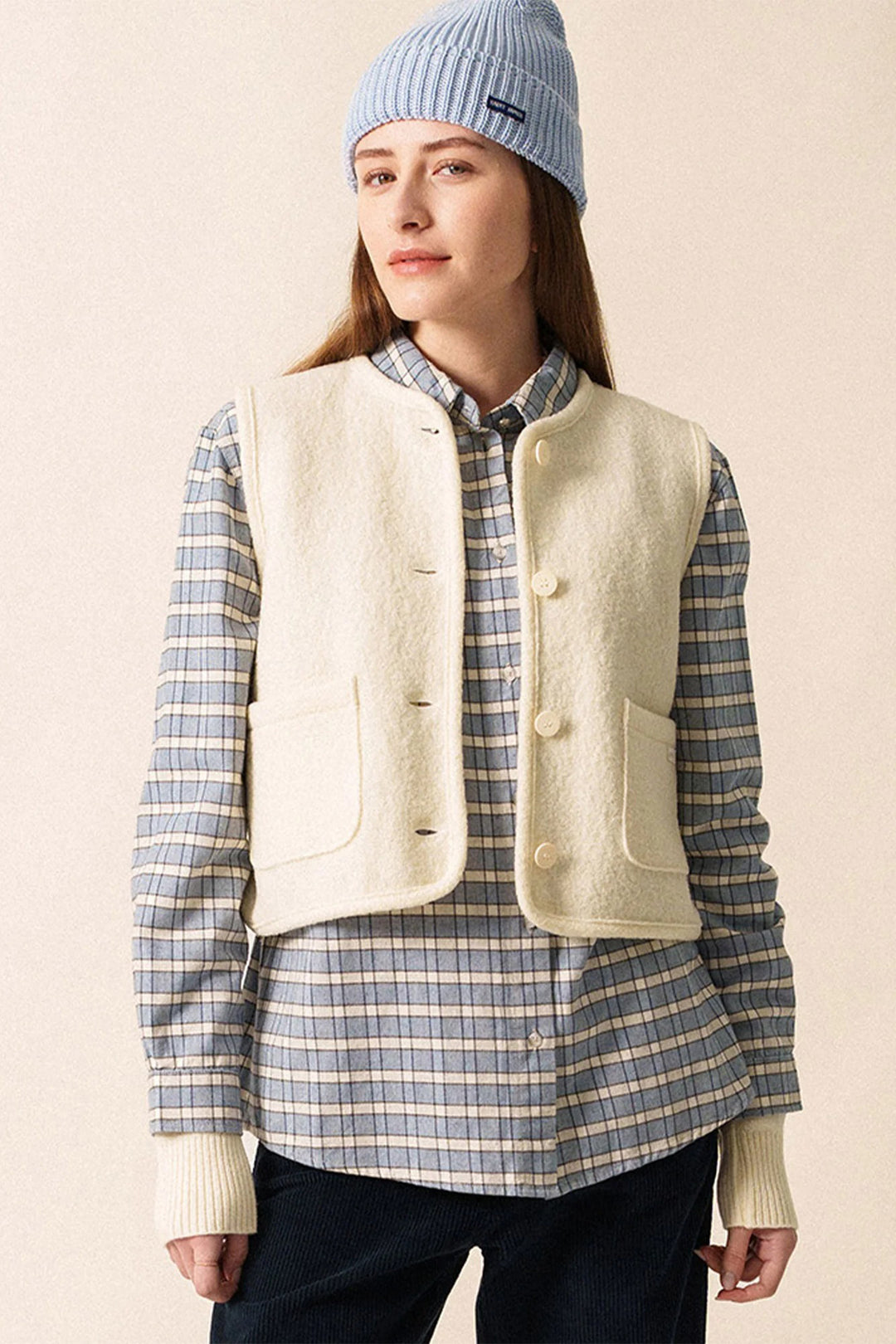 Person wearing a light-colored vest over a plaid shirt with a blue beanie on a beige background