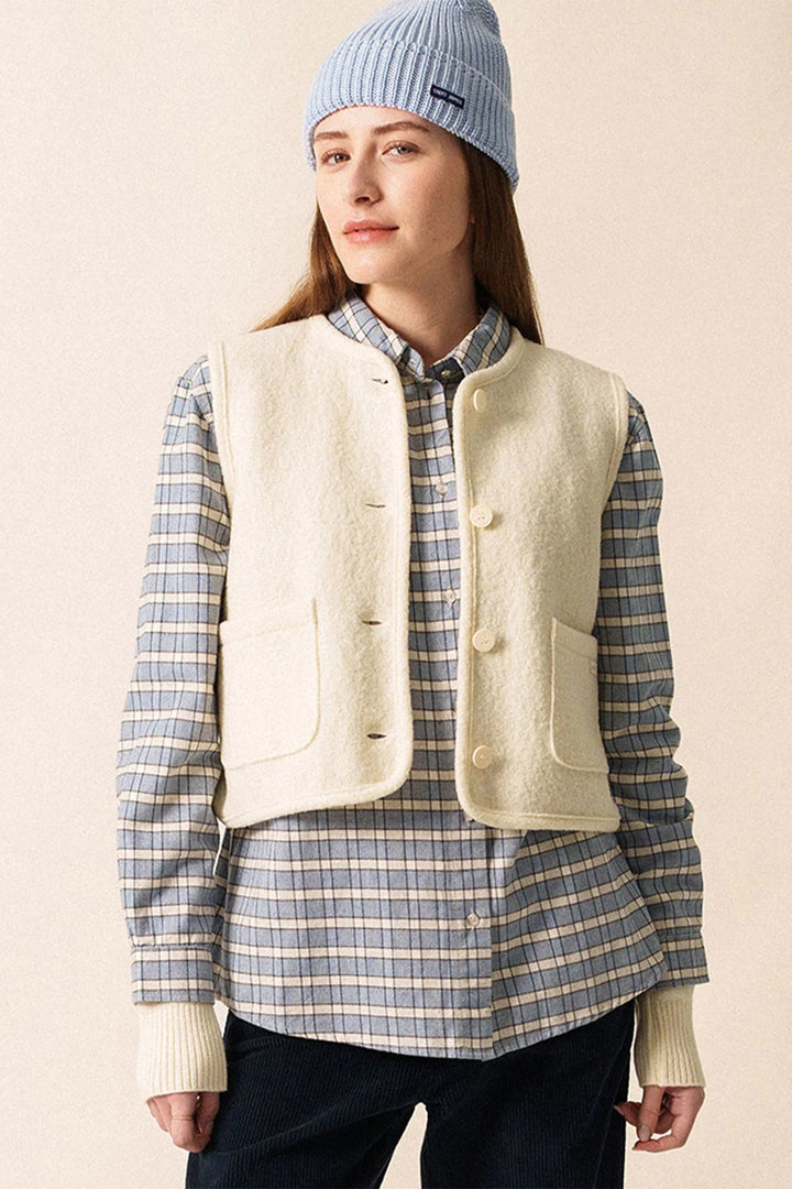 Person wearing a light-colored vest over a plaid shirt with a blue beanie on a beige background