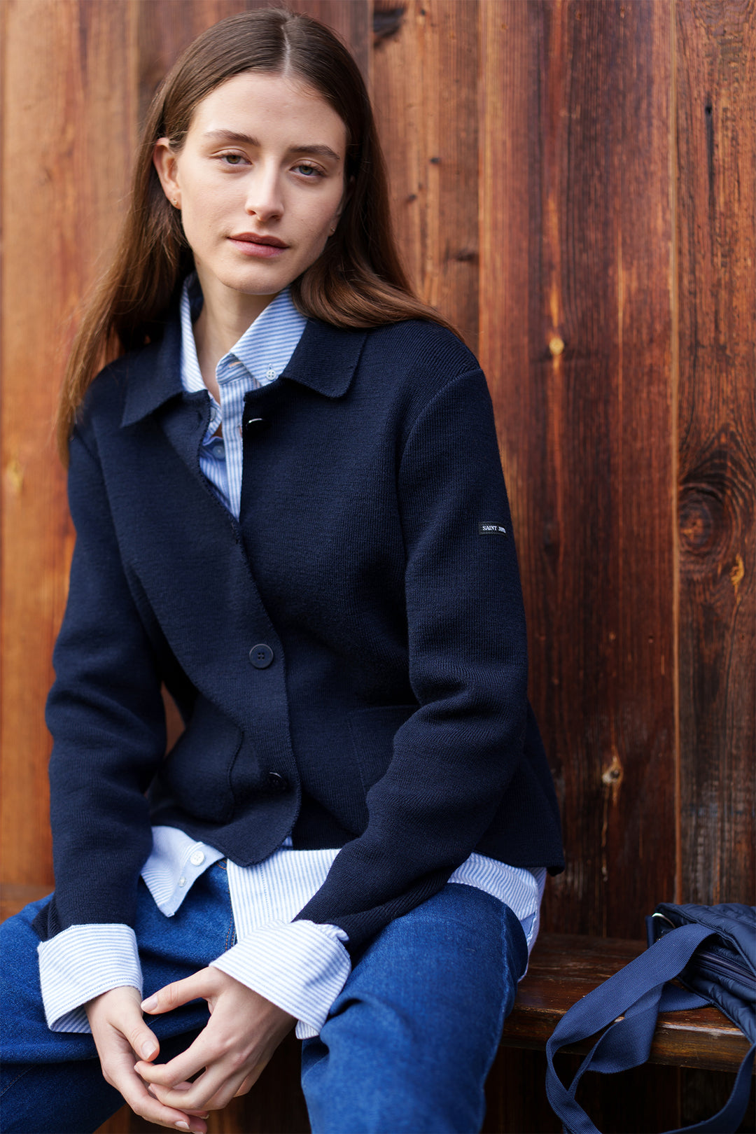 Woman wearing a navy coat and blue jeans sitting against a wooden wall.