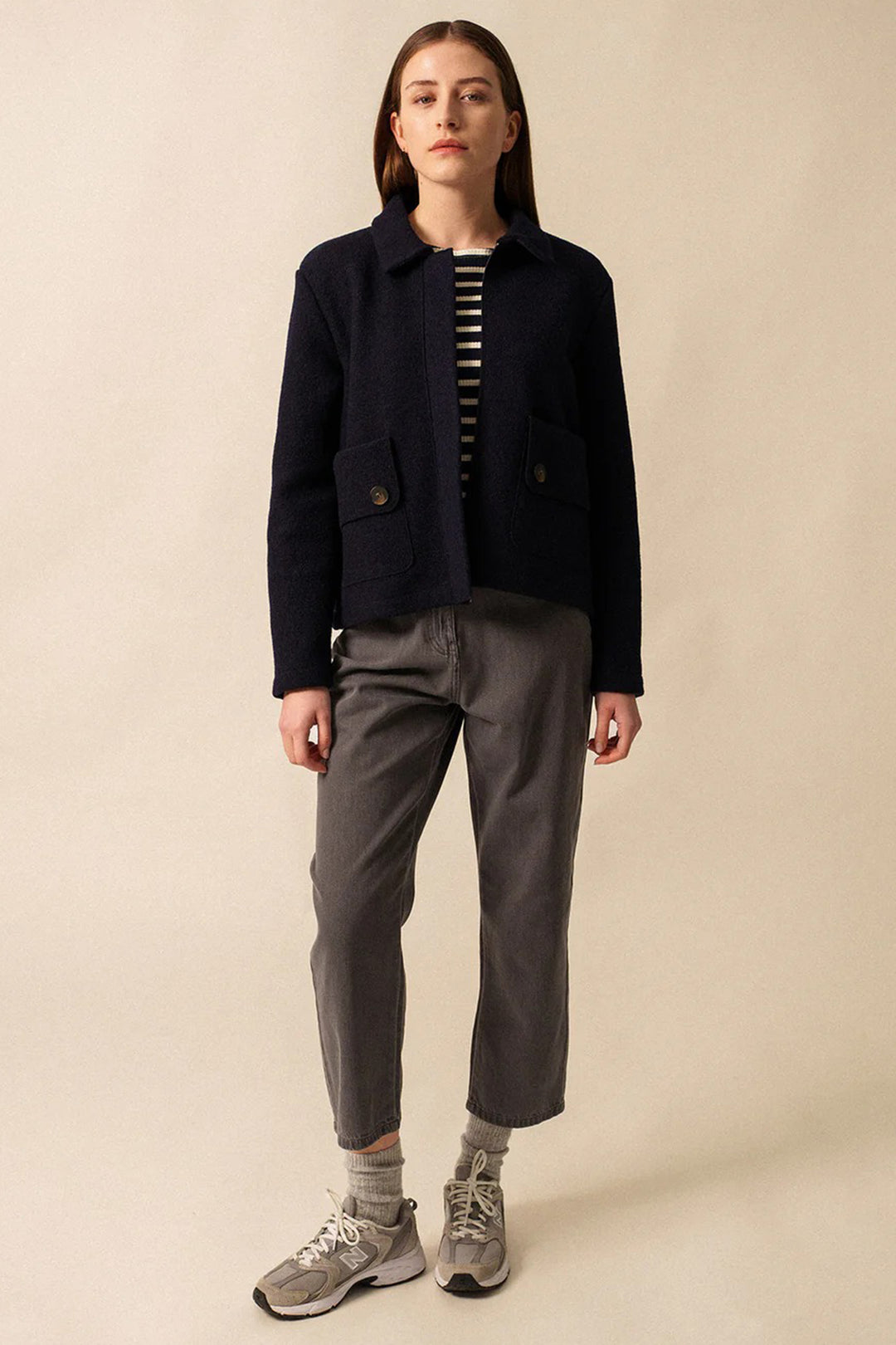 Person wearing a navy jacket, striped shirt, gray pants, and sneakers on a beige background
