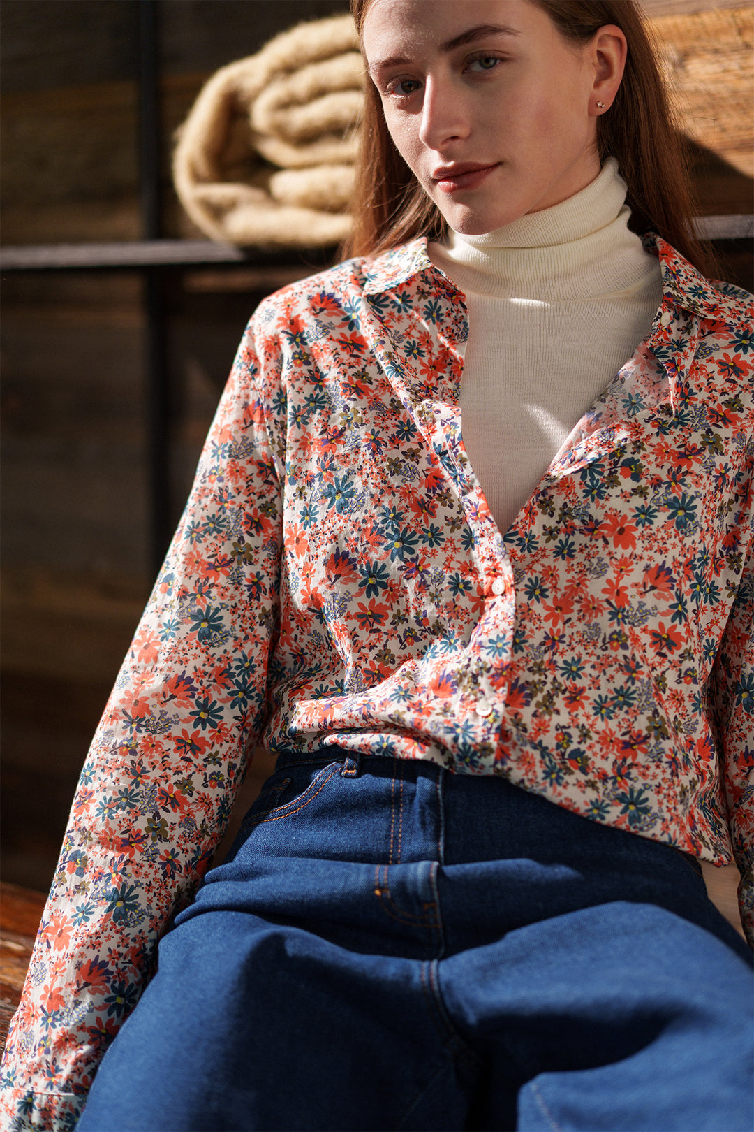 Woman wearing a floral blouse and blue jeans sitting indoors.