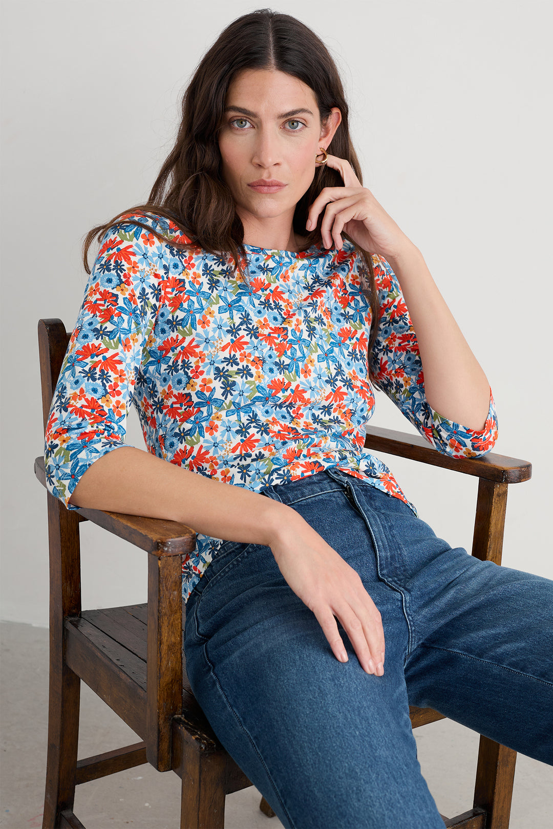 Woman sitting on a wooden chair wearing a colorful floral blouse and blue jeans.