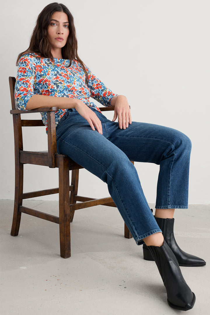 Woman sitting on a wooden chair wearing a floral top, blue jeans, and black boots.