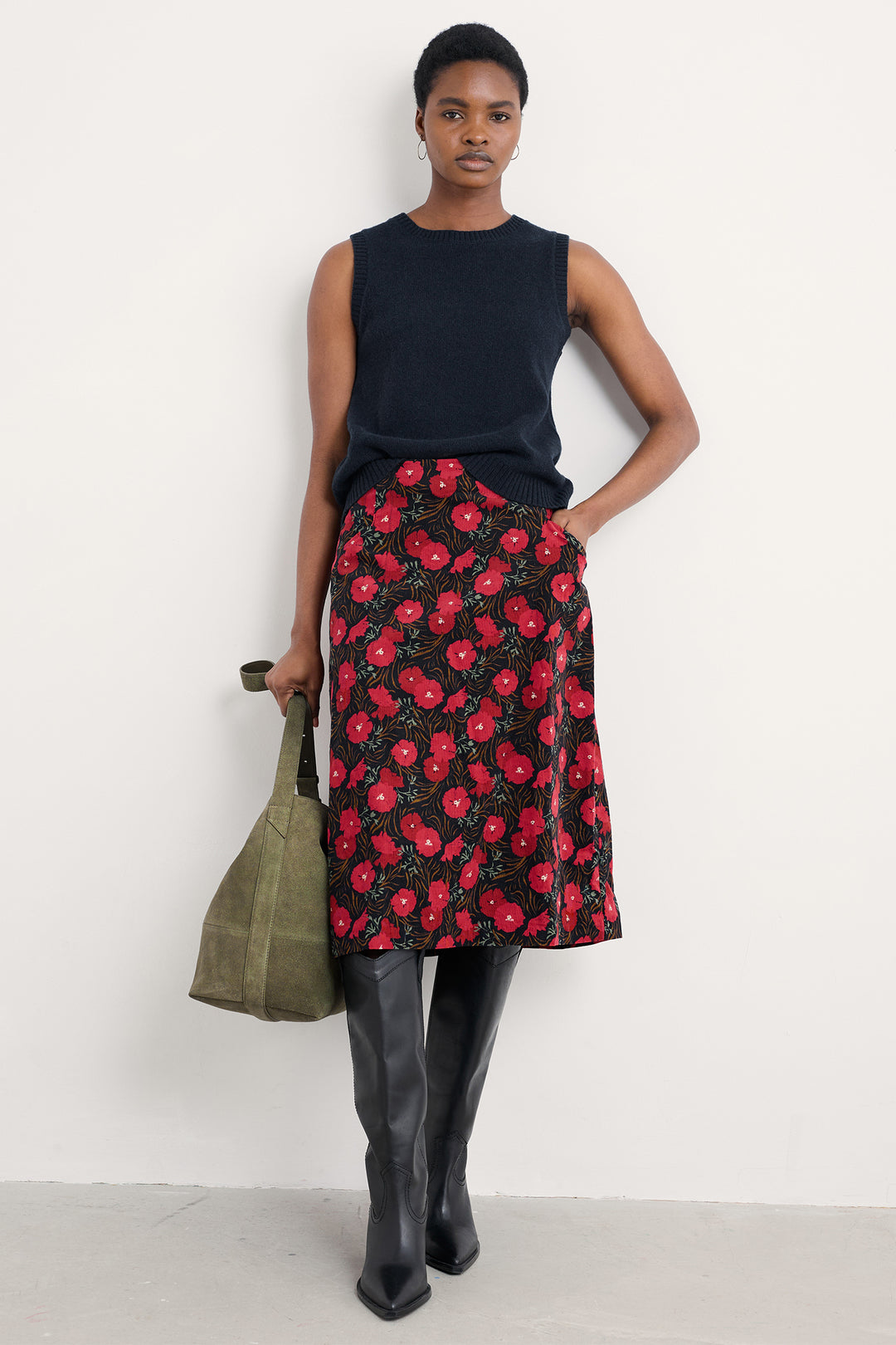 Woman wearing a black sleeveless top and red floral skirt, holding a green bag against a white background.