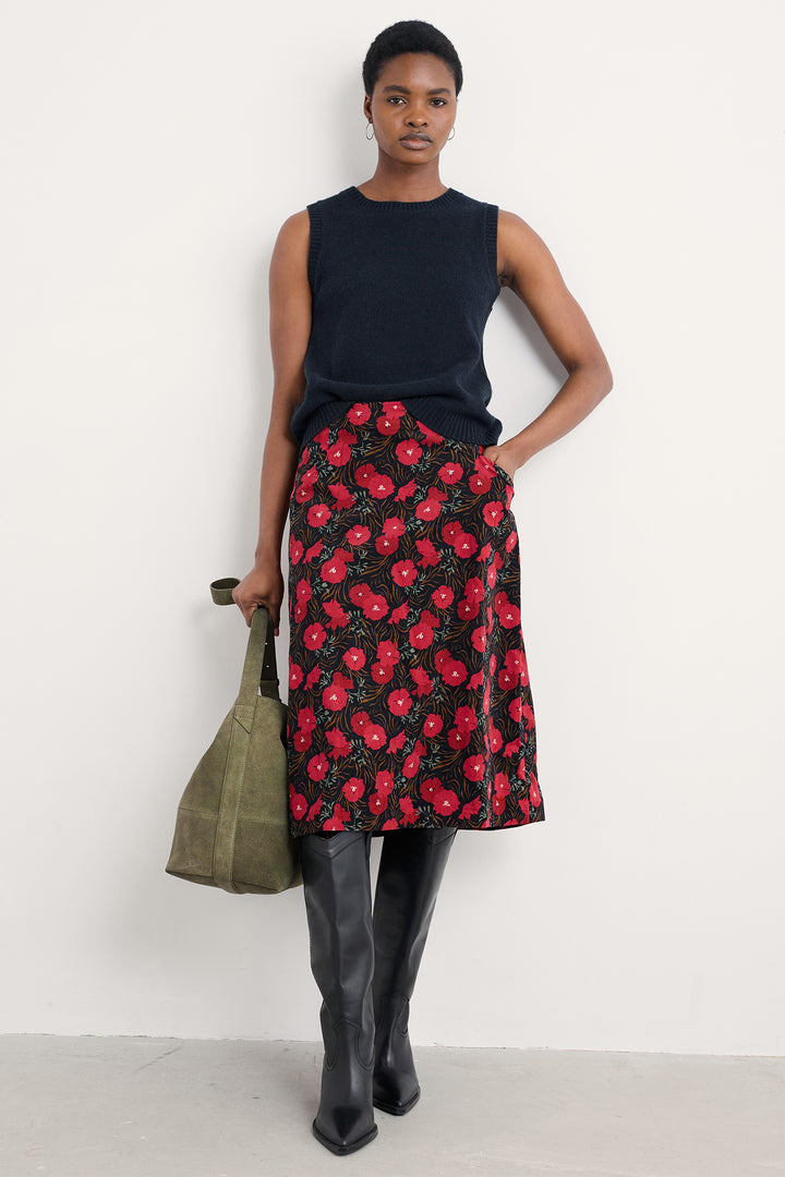Woman wearing a black sleeveless top and red floral skirt, holding a green bag against a white background.