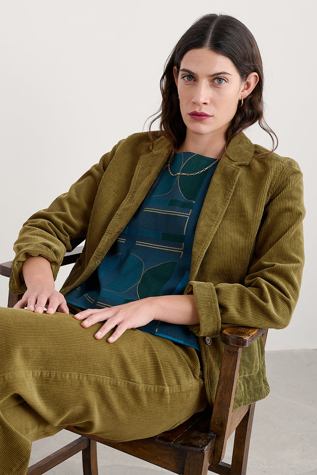 Woman wearing a green corduroy jacket sitting on a wooden chair against a white background