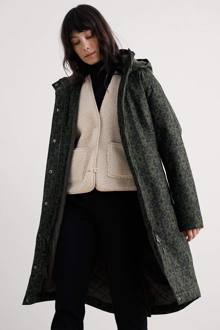 Person wearing a long dark green coat over a neutral sweater and black skirt on a white background