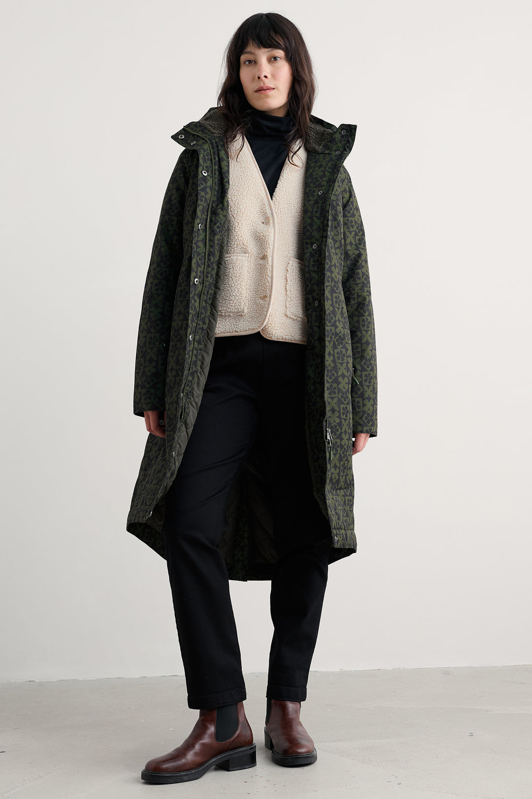 Person wearing a long green coat over a white sweater with black pants and shoes on a white background