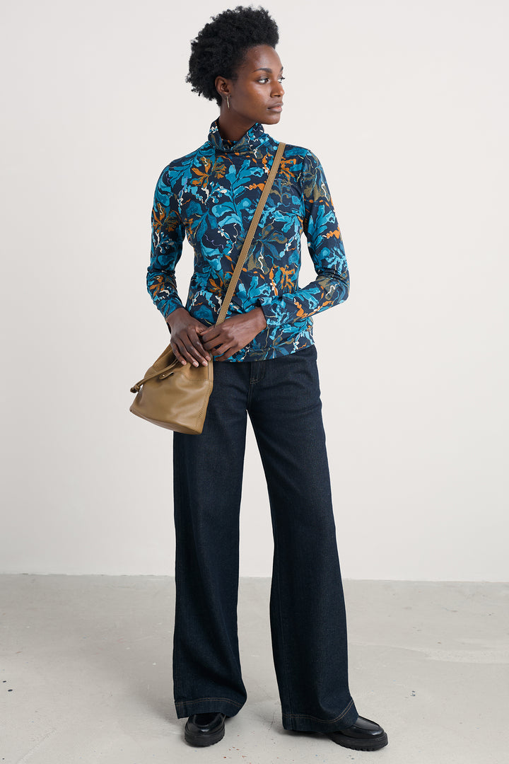 Woman wearing a blue patterned shirt and dark pants, holding a beige handbag on a white background
