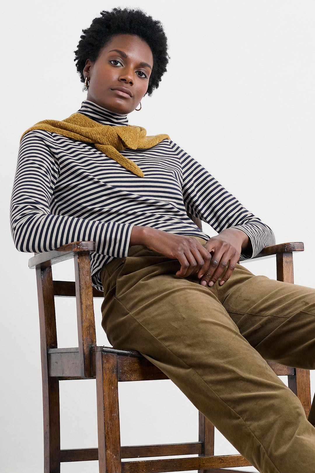 Person wearing a striped sweater and mustard yellow scarf sitting on a wooden chair against a white background
