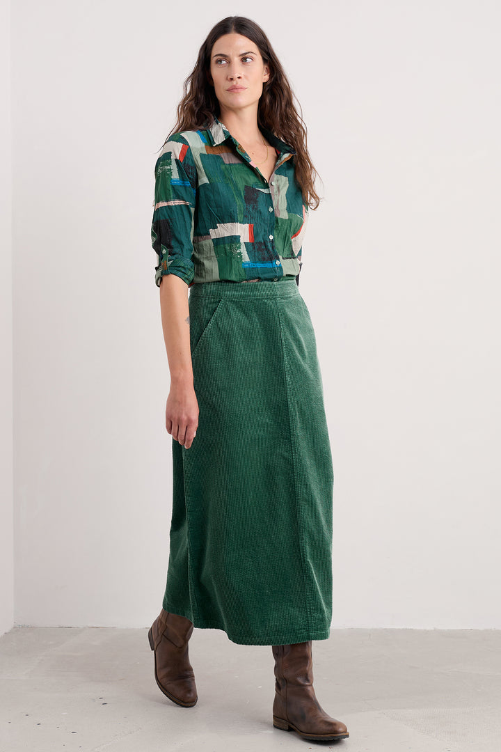 Woman wearing a green patterned shirt and green skirt with brown boots on a white background