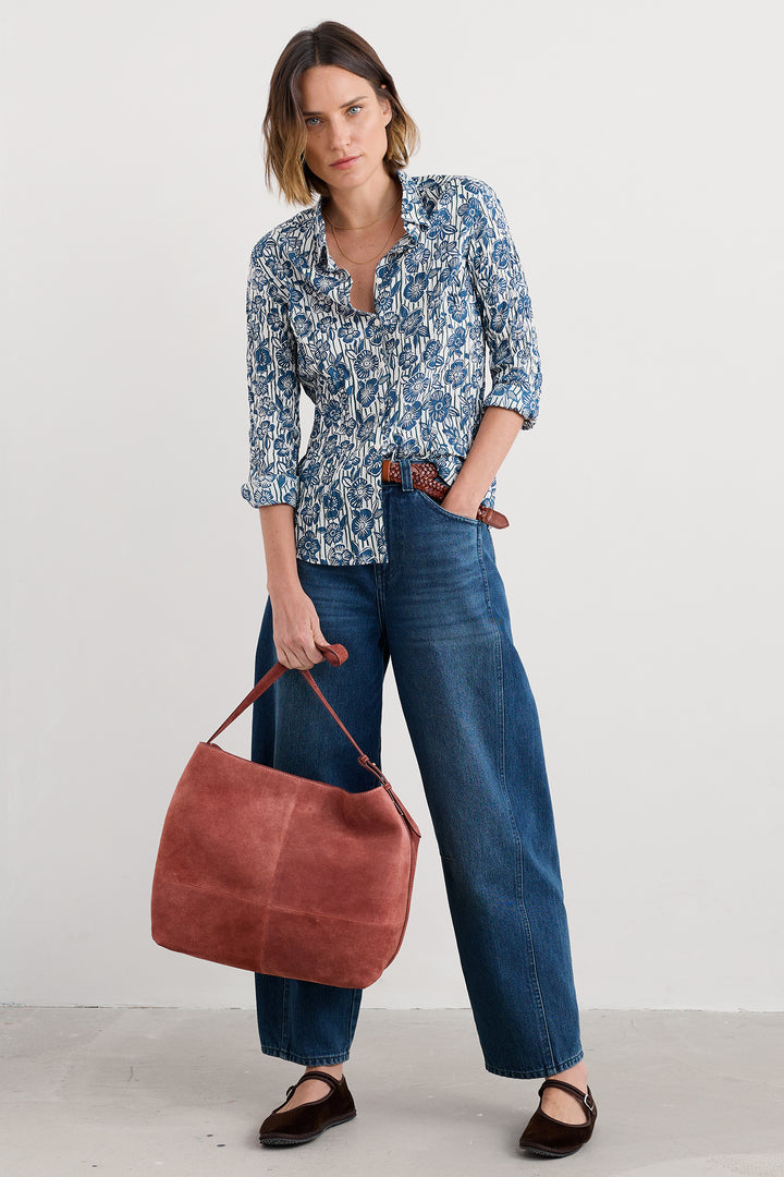 Woman wearing a blue patterned blouse and jeans, holding a brown leather bag against a white background