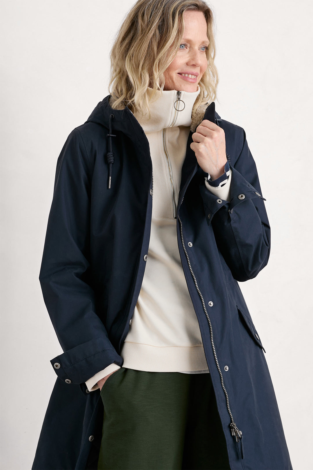 Woman wearing a navy blue coat over a white sweater and green pants on a light gray background