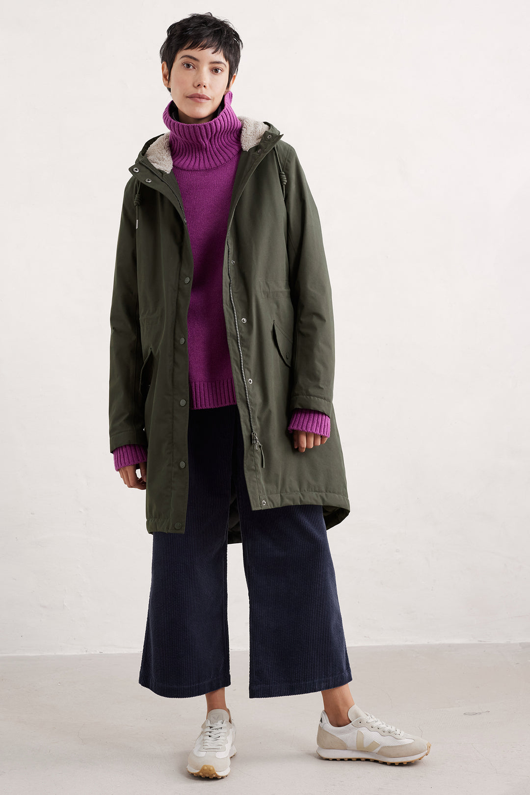 Person wearing a green coat, purple sweater, and dark pants on a white background