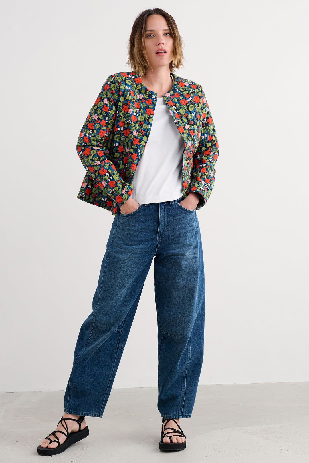 Woman wearing a colorful floral jacket, white shirt, blue jeans, and black sandals on a white background