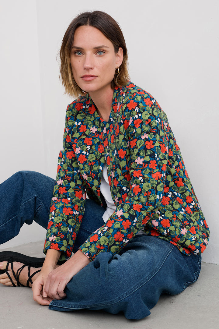 Woman wearing a colorful floral shirt and blue jeans sitting on the floor against a white wall.