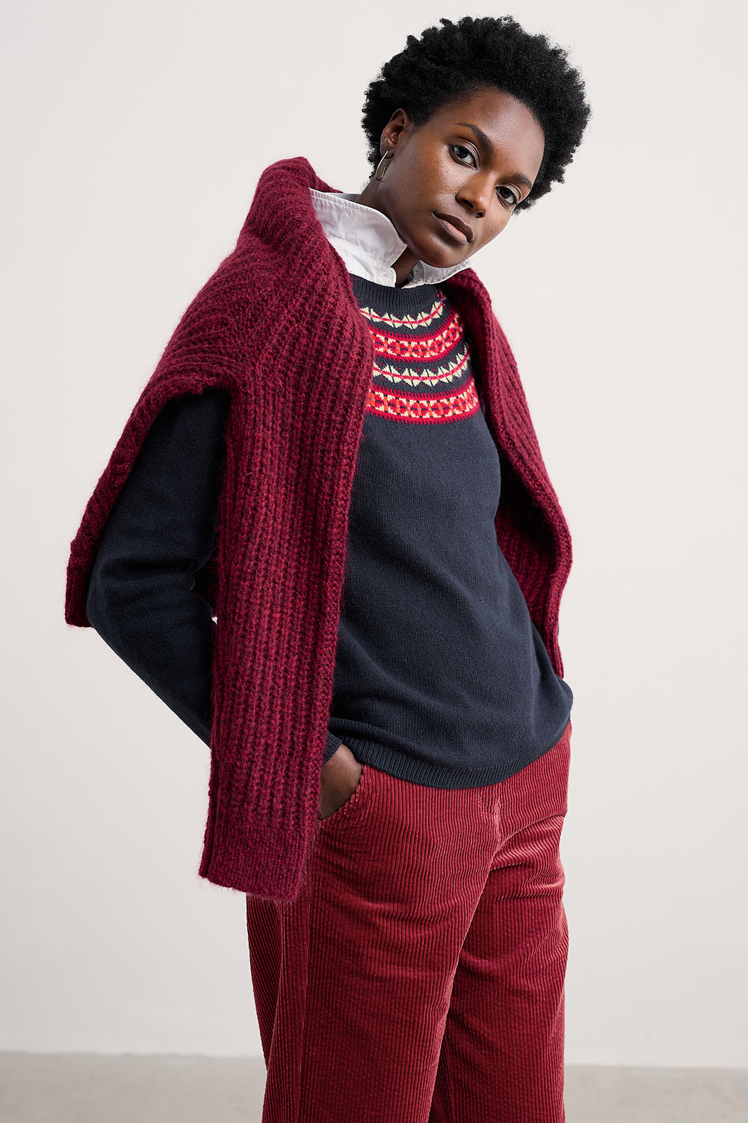 Person wearing a red knitted poncho over a navy sweater with a colorful pattern, paired with red pants against a plain background.