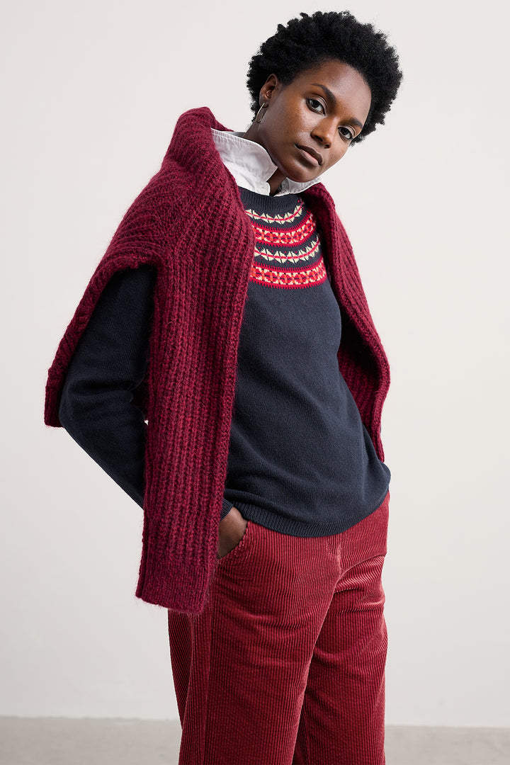 Person wearing a red knitted poncho over a navy sweater with a colorful pattern, paired with red pants against a plain background.