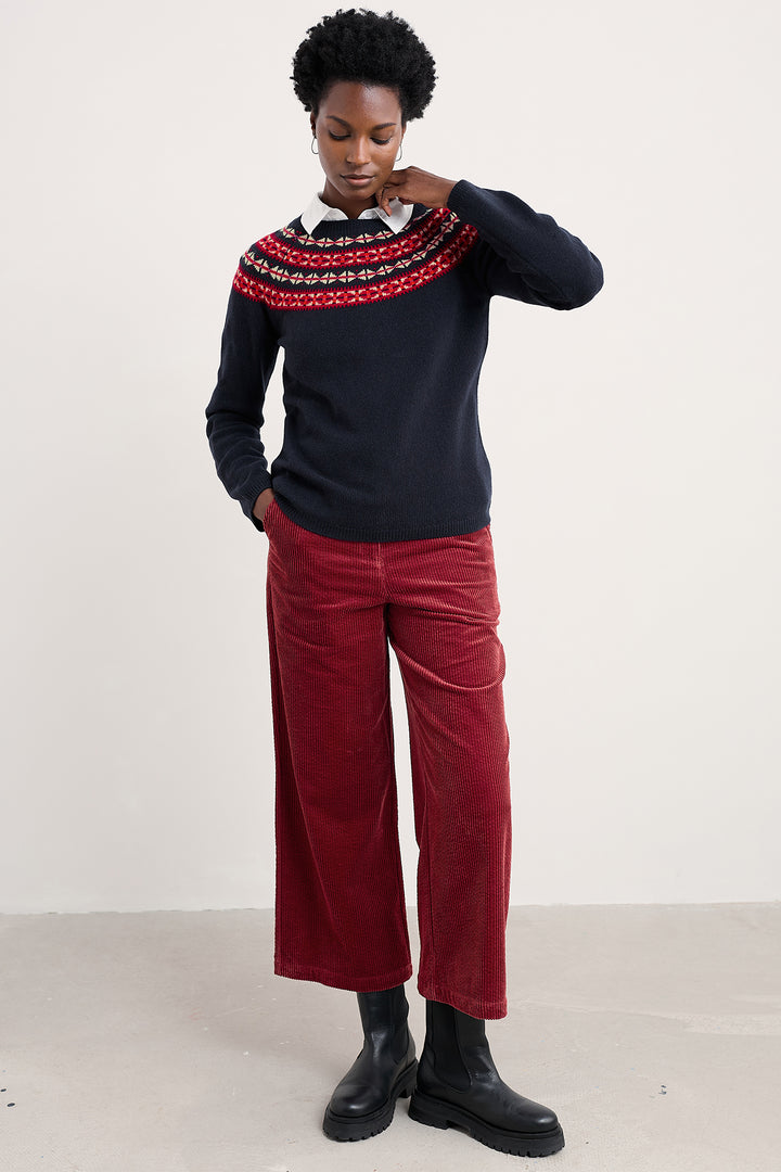 Person wearing a dark sweater with a colorful pattern, red pants, and black shoes on a white background