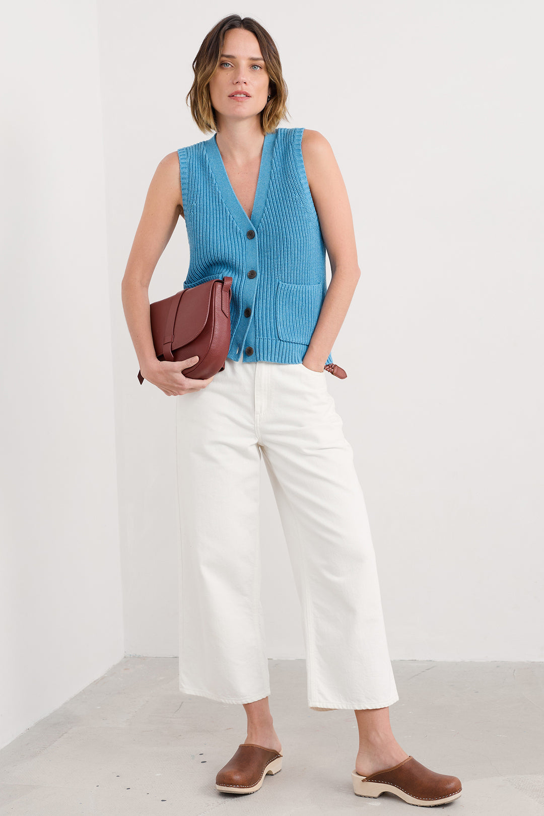 Woman wearing a blue sleeveless sweater vest, white pants, and brown shoes against a white background
