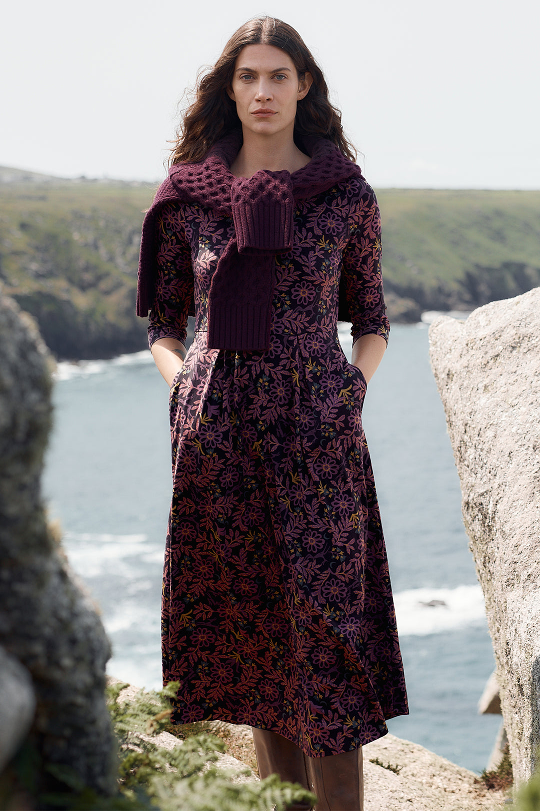 Woman in a dark floral dress standing by a body of water with cliffs in the background