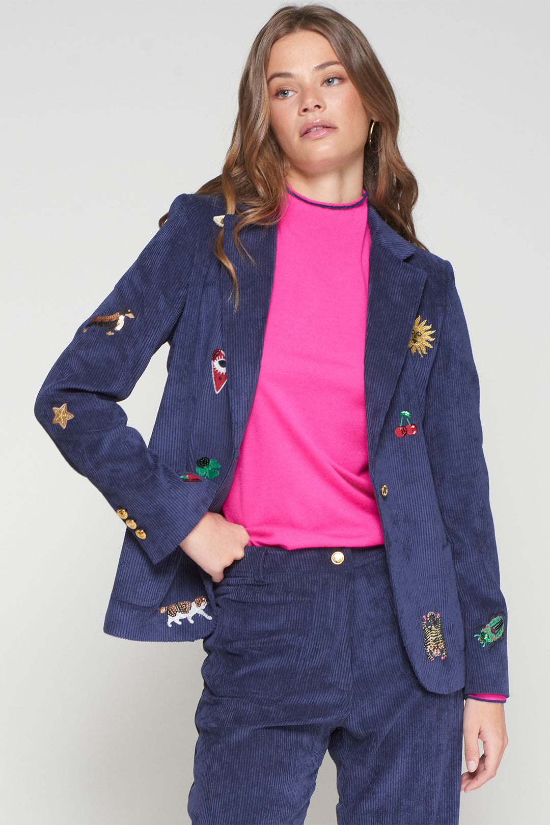 Woman wearing a navy blazer with colorful patches over a pink shirt and blue pants on a white background